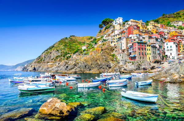 Busreisen ab Berlin nach Italien zur Cinque Terre. Riomaggiore in den Cinque Terre, Italien, mit bunten Häusern an der Felsküste und kleinen Booten im türkisblauen Wasser.