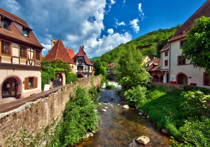 Busreisen nach Frankreich in das Elsass. Malerischer Blick auf Kaysersberg im Elsass, Frankreich, mit Fachwerkhäusern, Stadtmauer und Bachlauf vor grünen Hügeln.