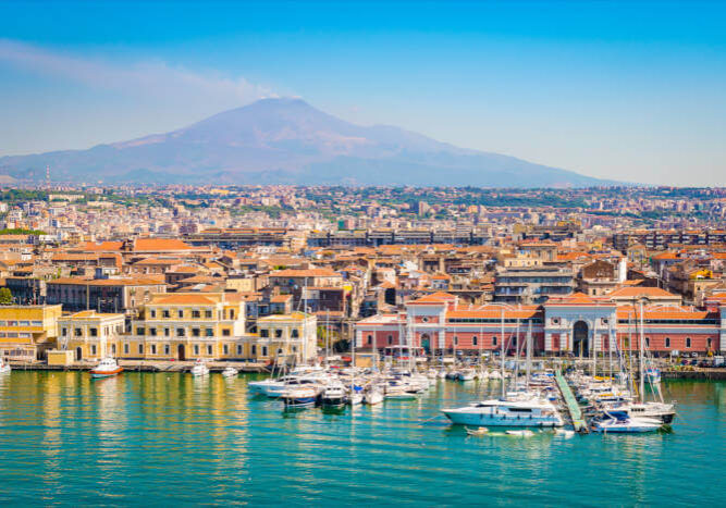 Hafen von Catania auf Sizilien, Italien, mit Segelbooten, historischen Gebäuden und dem Ätna im Hintergrund.