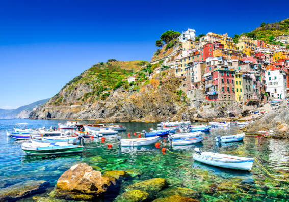 Busreisen ab Berlin nach Italien zur Cinque Terre. Riomaggiore in den Cinque Terre, Italien, mit bunten Häusern an der Felsküste und kleinen Booten im türkisblauen Wasser.
