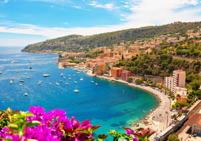 Cote dAzur Frankreich Reisen. Küstenblick auf die Côte d’Azur, Frankreich, mit türkisblauem Meer, Booten, bunten Häusern und blühenden Bougainvilleen im Vordergrund.