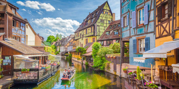Fachwerkhäuser am Kanal in der Altstadt von Colmar, Elsass, Frankreich, mit Boot und blühenden Balkonen bei sonnigem Wetter.