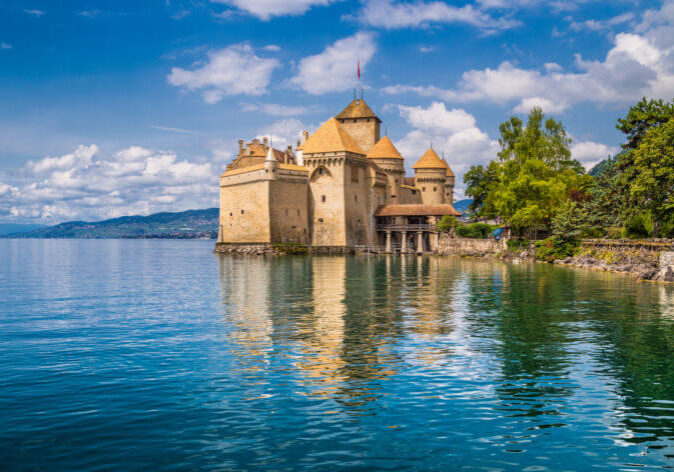 Reise Inspirationen Schweiz Reisen mit Blick auf Genf. Schloss Chillon am Ufer des Genfersees, Schweiz, mit Spiegelung im Wasser und Alpenpanorama im Hintergrund.