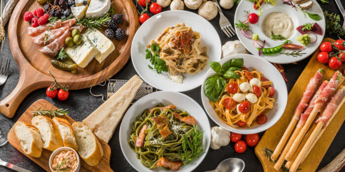 Auswahl italienischer Spezialitäten mit Pasta, Käse, Antipasti, Brot und Schinken auf rustikalem Holztisch.